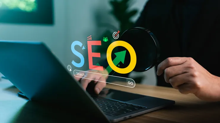 Laptop on a desk with colorful SEO letters and a magnifying glass, illustrating search engine optimization.