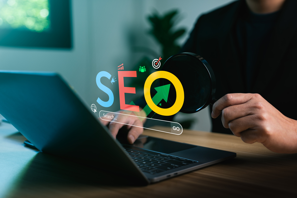 Laptop on a desk with colorful SEO letters and a magnifying glass, illustrating search engine optimization.